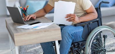 Paraplegic black guy in wheelchair with documents using laptop to work online from home. Handicapped young Afro freelancer with papers making business project on internet