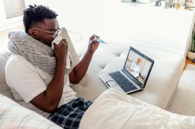 Sick young man blowing his nose talking to a doctor. Sick young male complaining during video call with his consulting physician. Sick man in pajamas covered with blanket sitting on sofa in living room and talking with his doctor online.