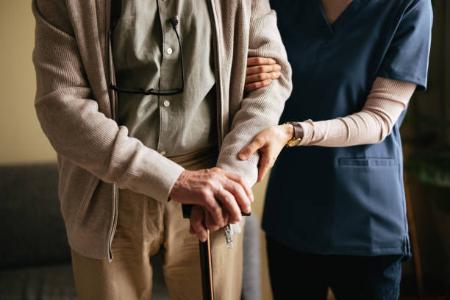 An anonymous Japanese female caregiver supporting elderly patient to stand and walk with a walking cane at the nursing home.